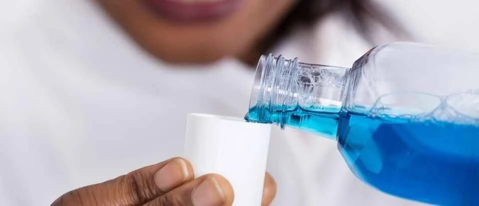 Una persona vierte enjuague bucal de color azul desde su botella hacia un pequeño vaso blanco.