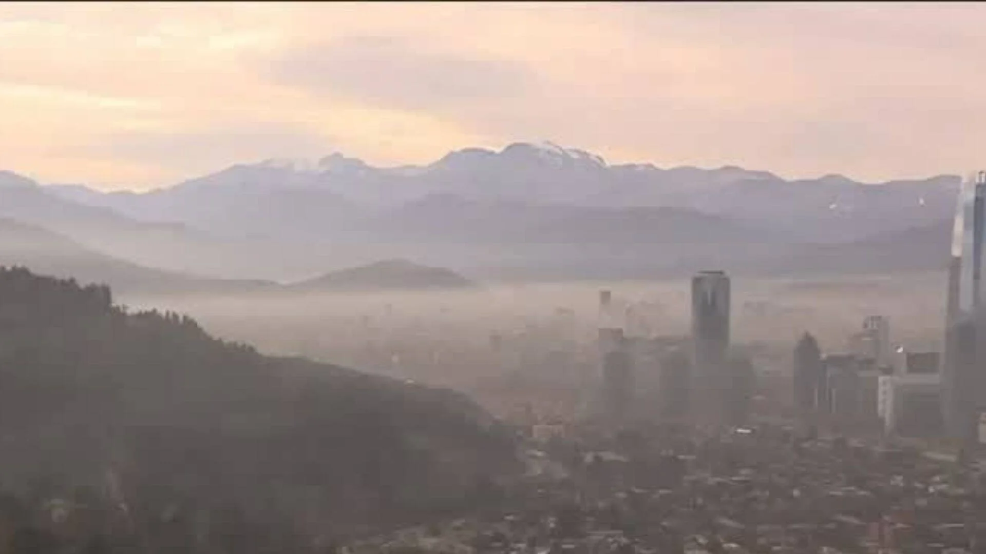 Vista aérea de una ciudad con densa capa de smog entre sus edificios y una cordillera nevada al fondo bajo cielo rosado.