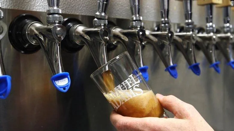 Mano sosteniendo un vaso de Samuel Adams mientras se sirve cerveza de barril en una fila de grifos metálicos.