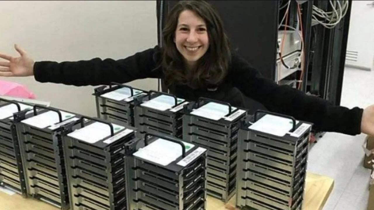 Mujer con los brazos abiertos posa orgullosa detrás de varias pilas de discos duros apilados sobre una mesa.
