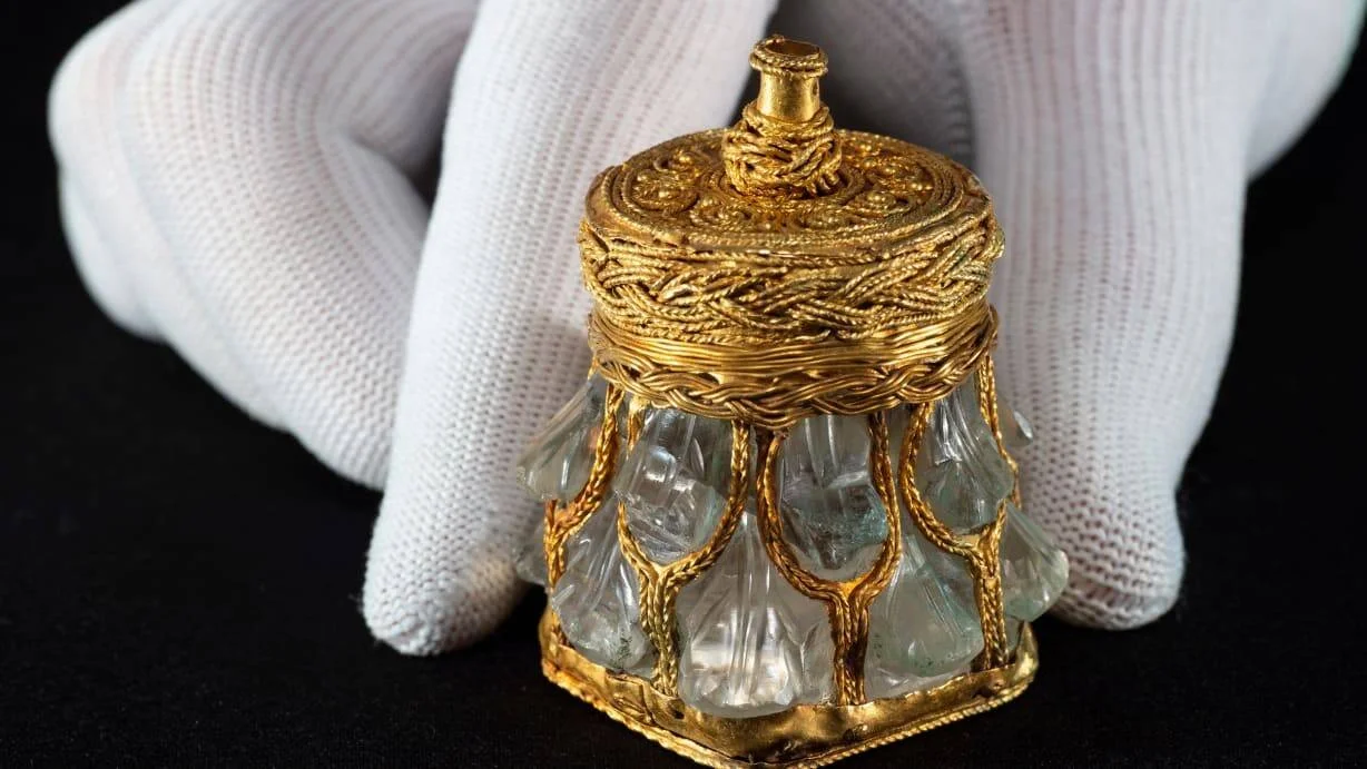 Manos con guantes blancos sostienen un pequeño cofre o relicario medieval decorado con filigrana de oro y paneles de cristal tallado.