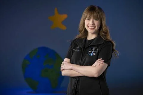 Mujer joven con traje negro de SpaceX posa con brazos cruzados ante un fondo con globo terráqueo y estrella pintados.