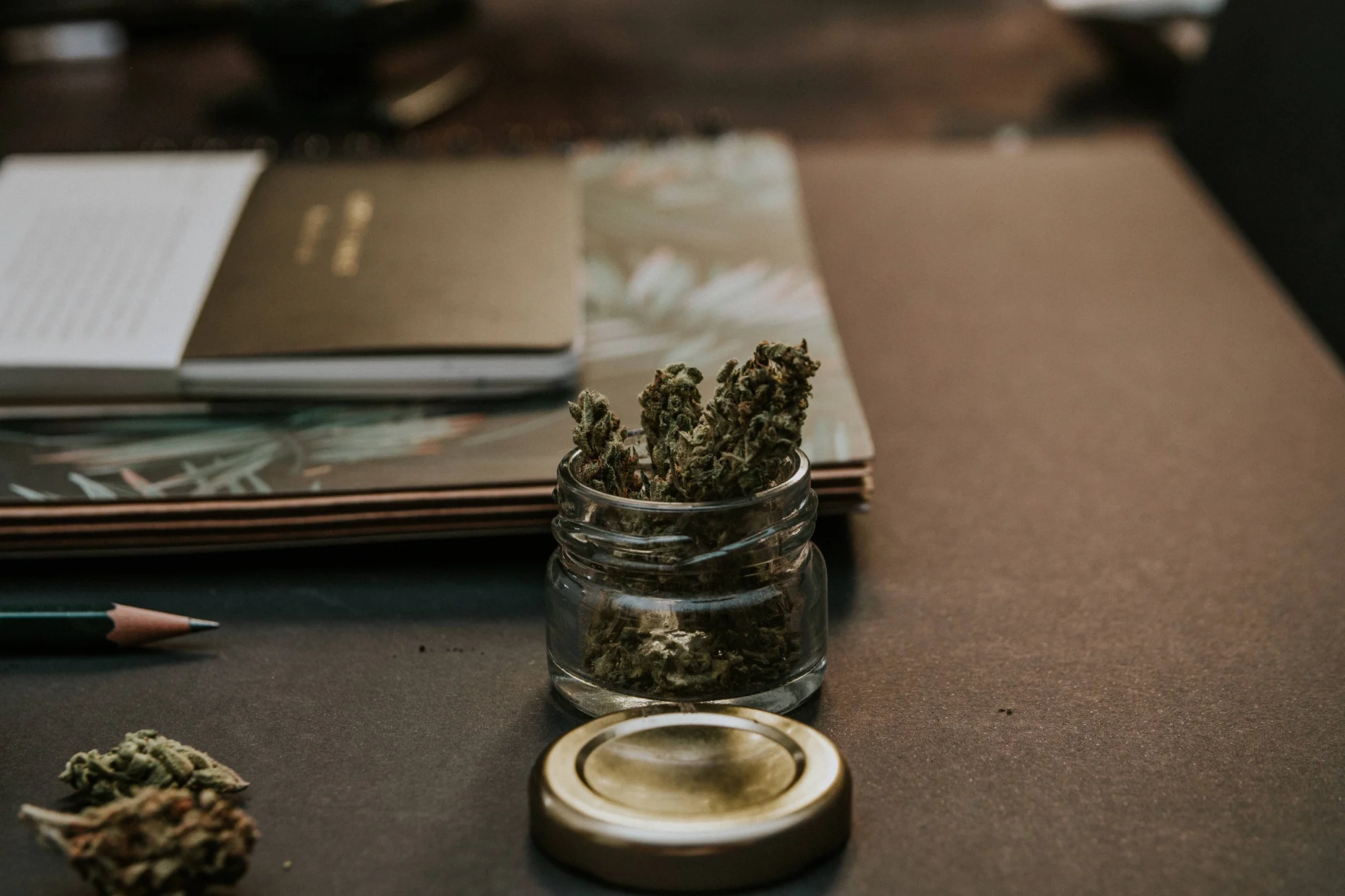 Frasco de vidrio abierto con cogollos de cannabis sobre una mesa junto a libros, un lápiz y la tapa dorada.