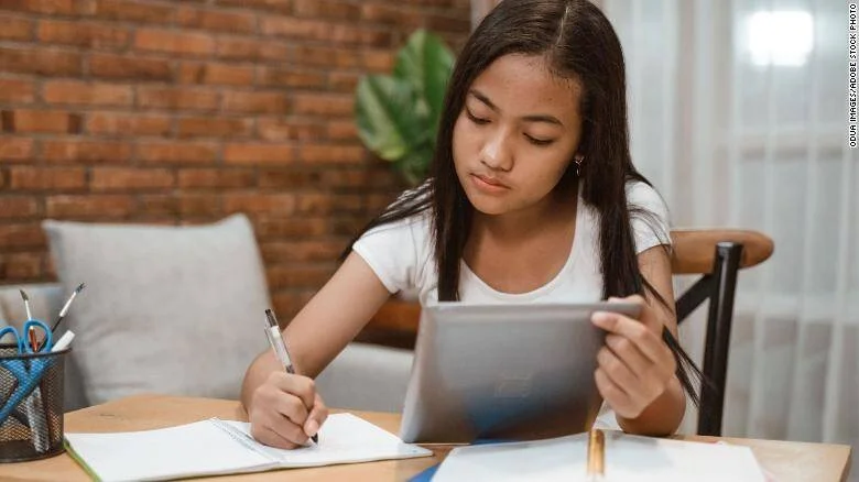 Una niña escribe en un cuaderno mientras sostiene una tablet, estudiando en un escritorio en casa.