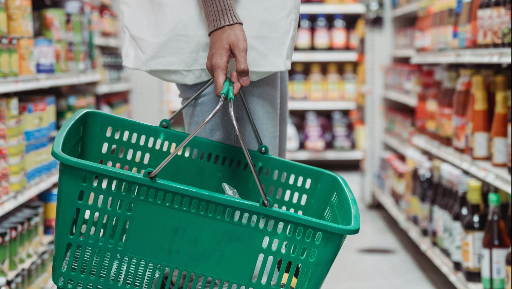 Una persona sostiene una canasta de compras verde vacía mientras recorre el pasillo de un supermercado.
