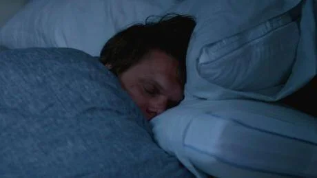 Una persona duerme profundamente cubierta con una frazada azul en un cuarto oscuro.
