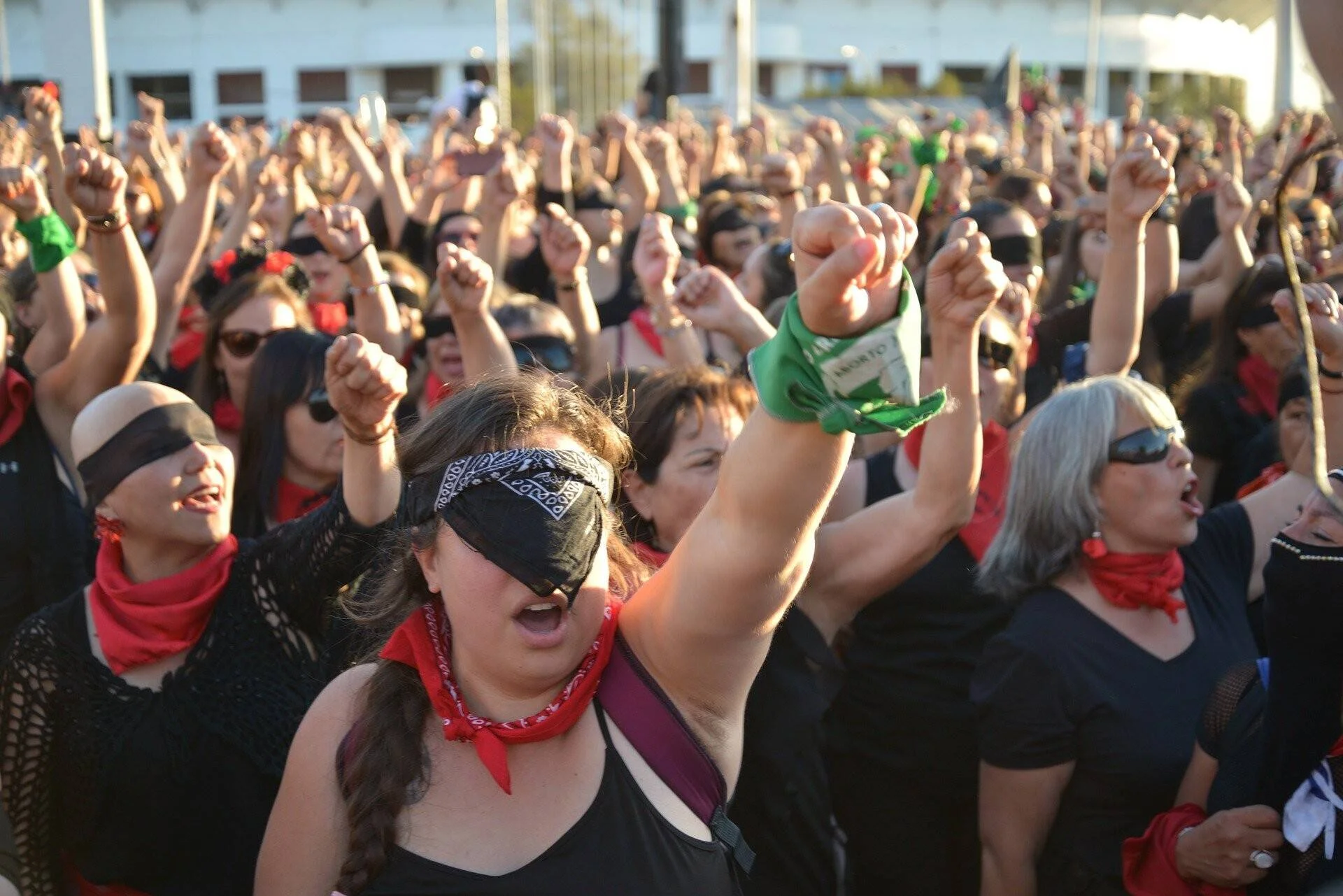 Multitud de mujeres vestidas de negro con vendas en los ojos y pañuelos rojos levantan los puños en una manifestación feminista.