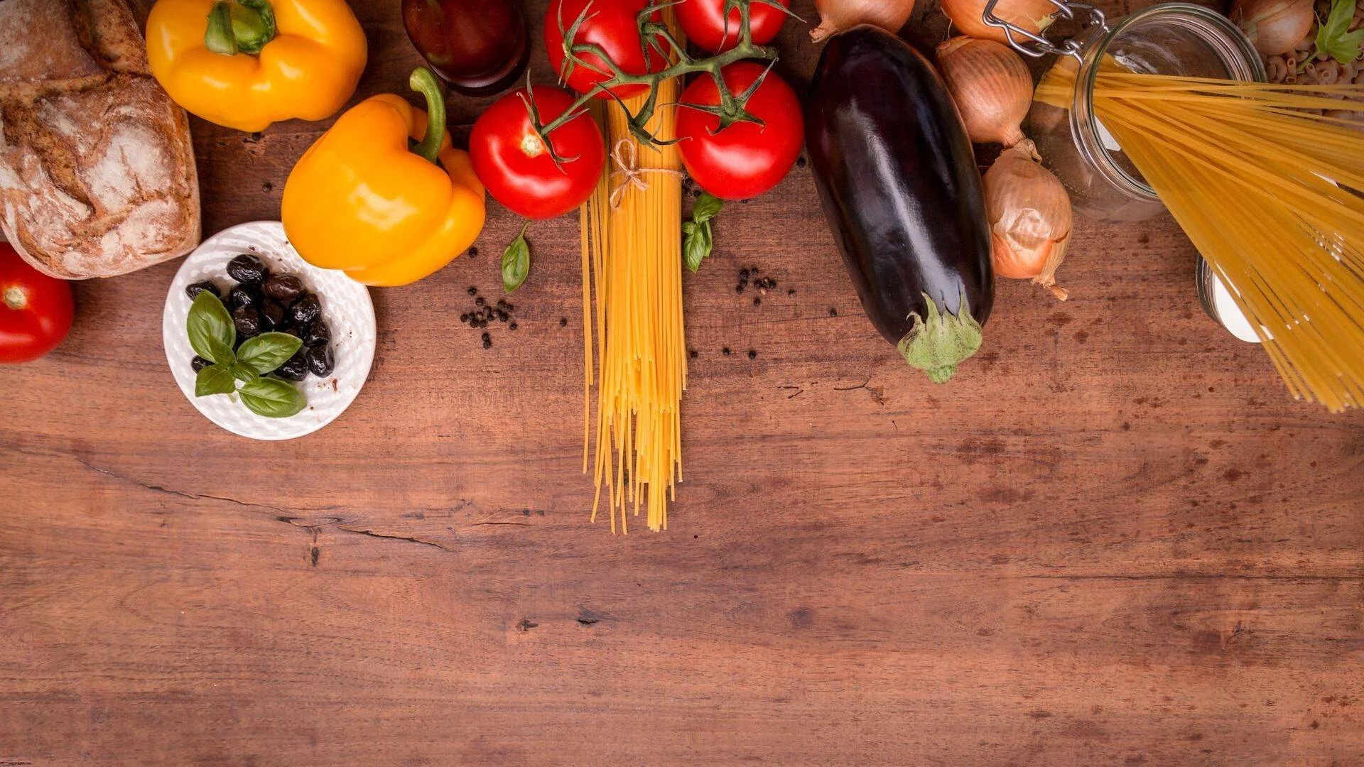 Tomates, pimientos, berenjena, cebollas, espaguetis crudos, aceitunas, pan y albahaca dispuestos sobre una mesa de madera.
