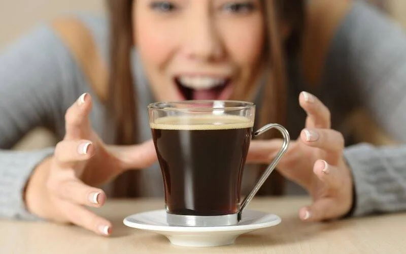 Mujer con expresión entusiasta estira las manos hacia una taza de café negro sobre una mesa.