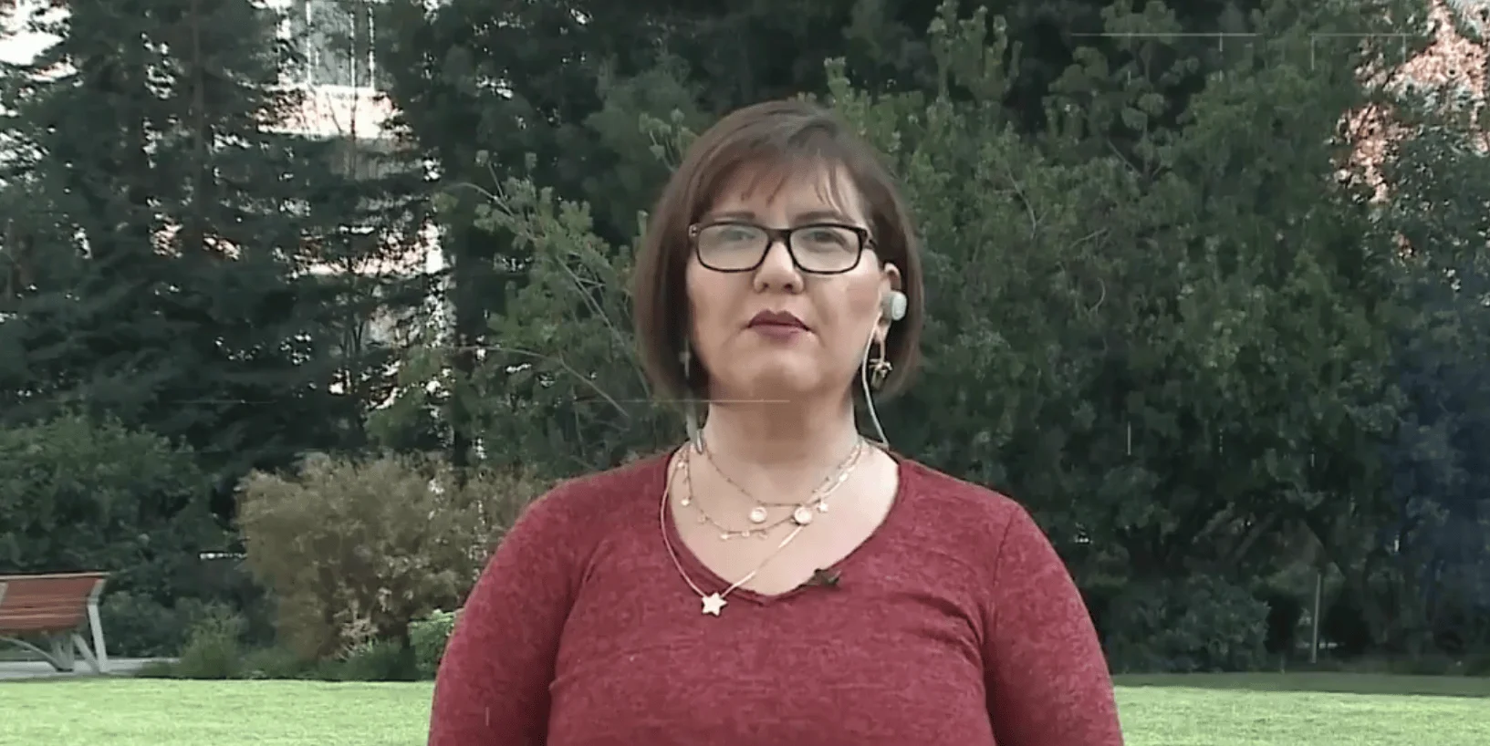 Mujer con gafas y auricular de periodista grabando una crónica en un parque con árboles de fondo.