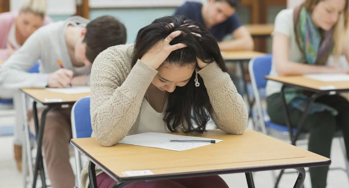 Joven estudiante con las manos en la cabeza, visiblemente estresada frente a un examen escrito en un aula con otros estudiantes.