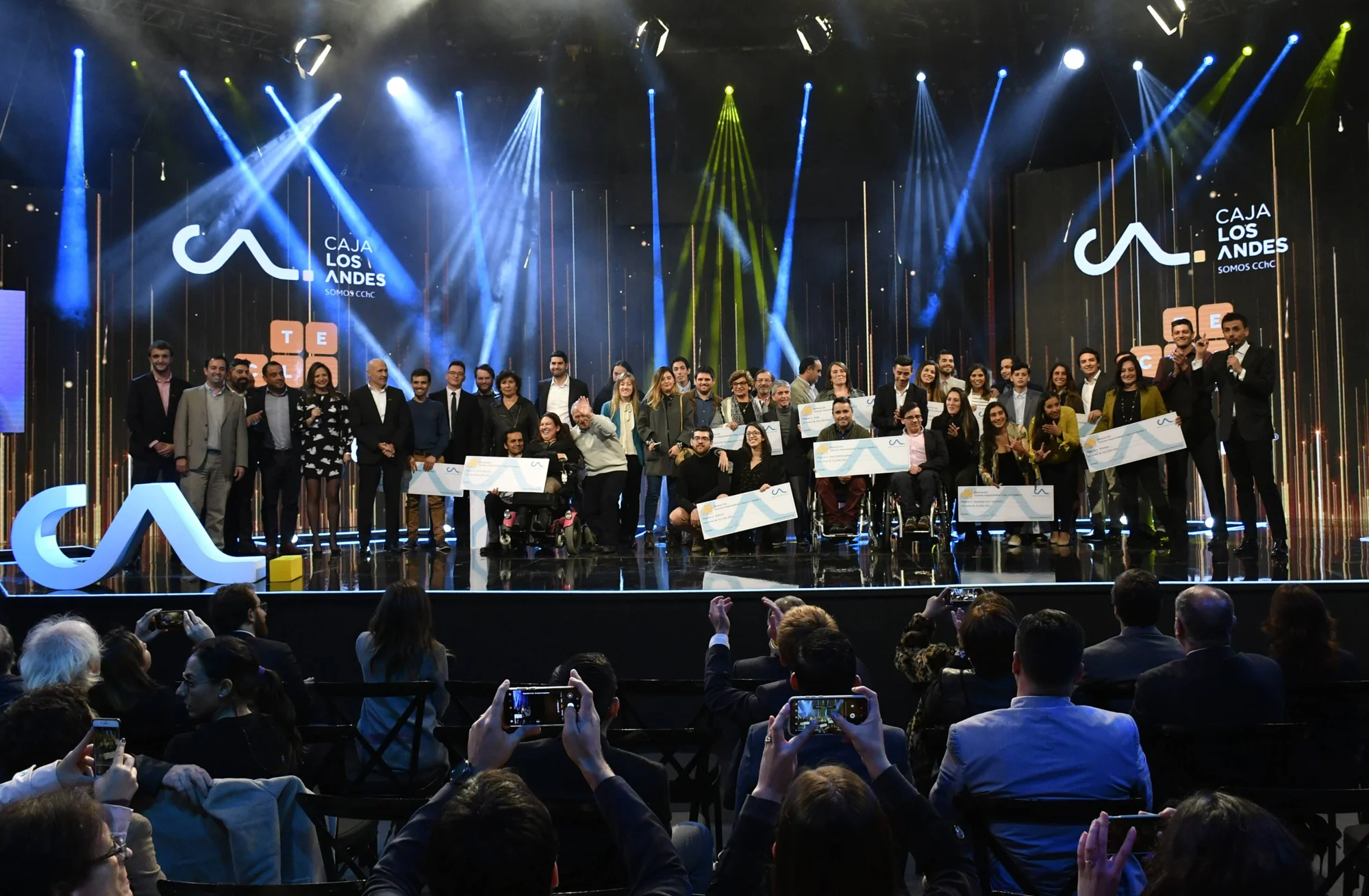 Grupo de ganadores sostiene cheques simbólicos en el escenario de la ceremonia de premiación de Caja Los Andes, ante un público que fotografía el momento.