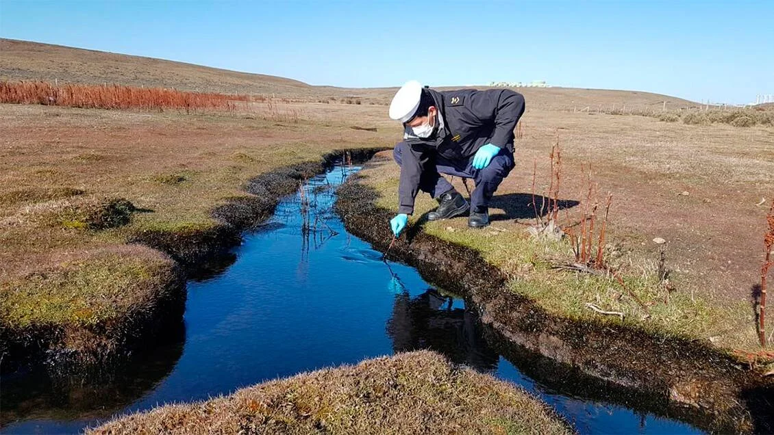 Un técnico con uniforme, mascarilla y guantes azules toma una muestra de agua en un pequeño arroyo en una llanura patagónica.