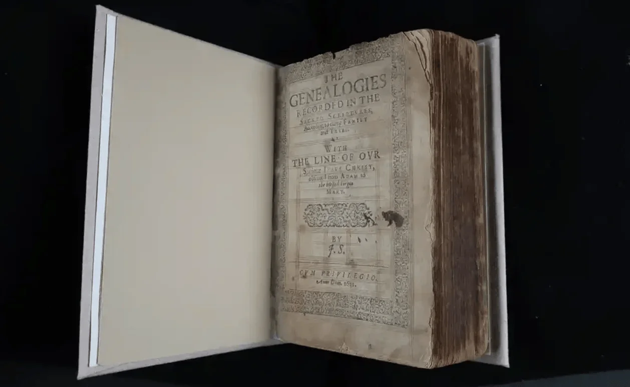 Libro antiguo abierto mostrando la portada de 'The Genealogies Recorded in the Sacred Scriptures', firmado por J.S., con sello Cum Privilegio.