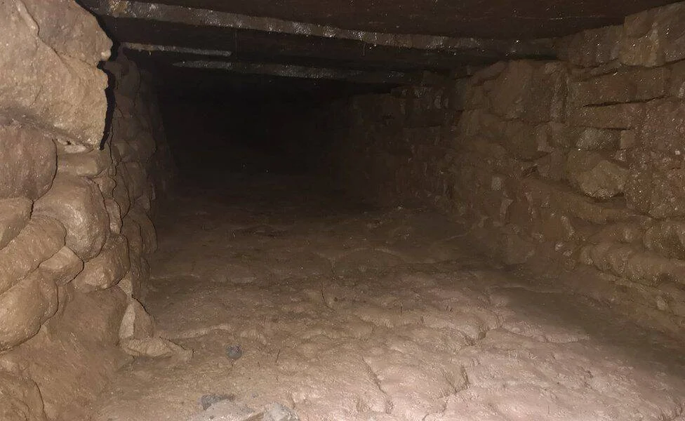 Interior de un túnel o cámara subterránea con paredes y suelo de piedra rugosa, techo bajo de madera y oscuridad al fondo.