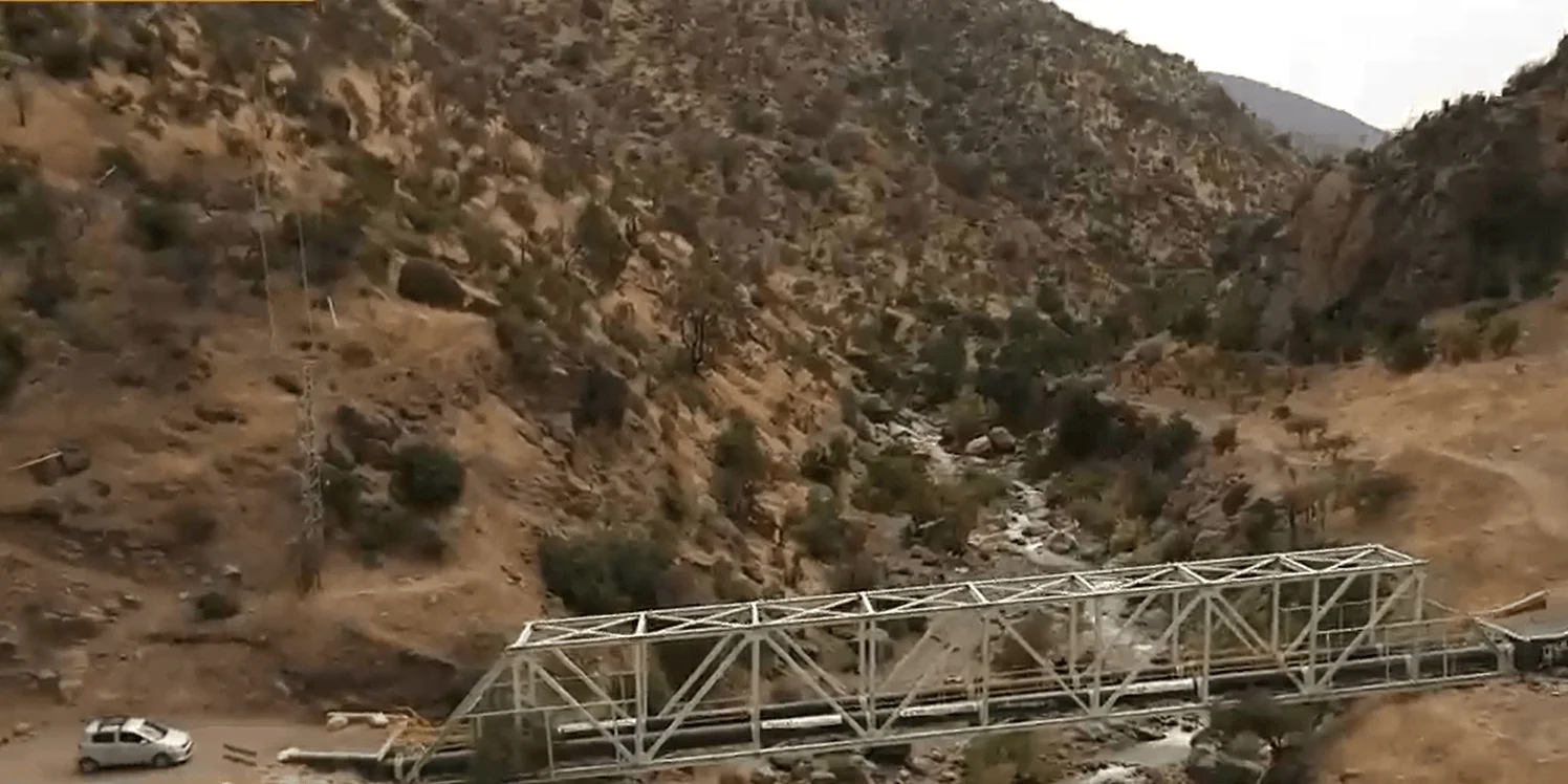 Puente de estructura metálica tipo celosía cruza una quebrada en un valle árido con cerros secos y un arroyo al fondo.