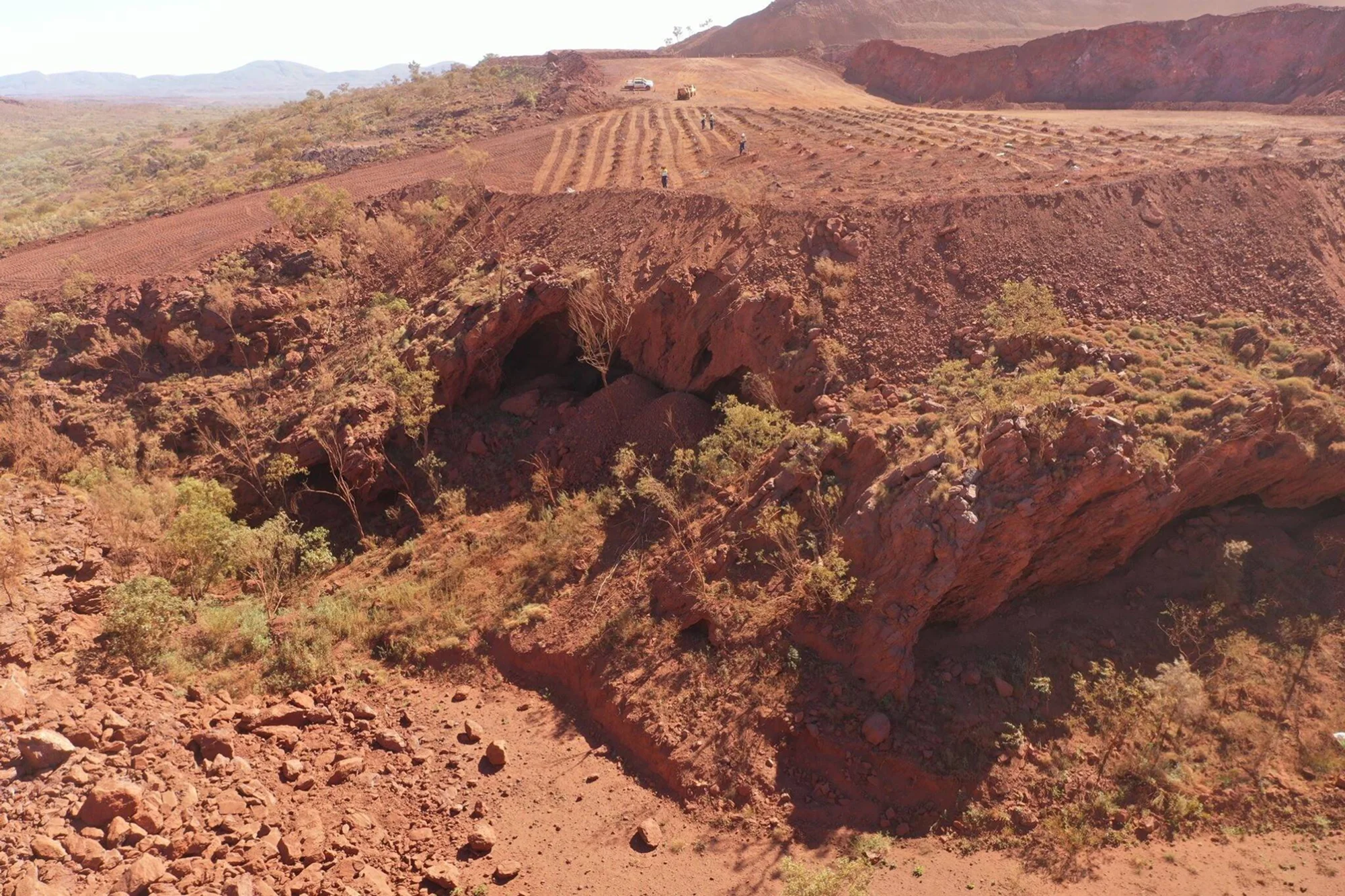Formación rocosa con cueva en terreno árido rojizo, con maquinaria y trabajadores en explanada minera al fondo.