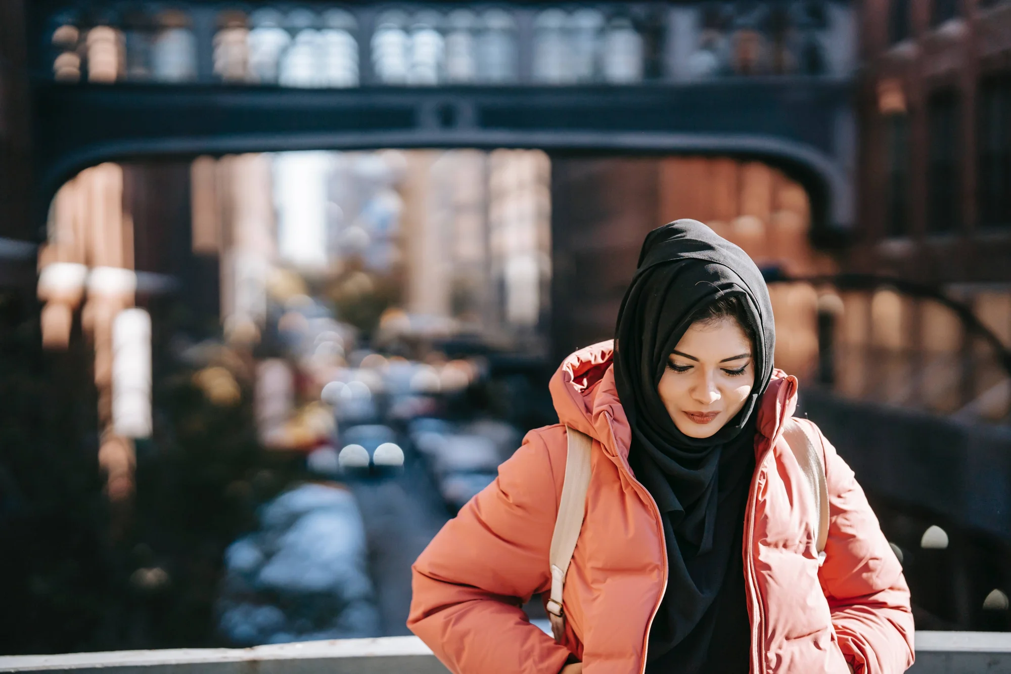 Mujer joven con hiyab negro y campera rosa apoyada en barandal urbano, con calle y puente desenfocados al fondo.