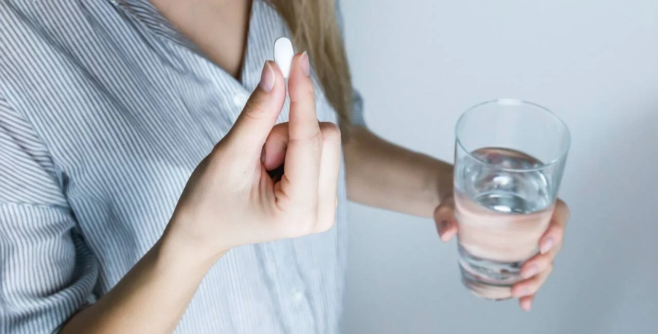 Mujer sostiene una pastilla blanca en una mano y un vaso de agua en la otra, a punto de tomarla.
