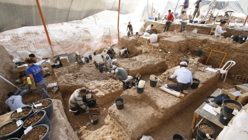 Numerosos arqueólogos trabajan en una amplia excavación cubierta por lonas, removiendo tierra con herramientas manuales y cubetas.