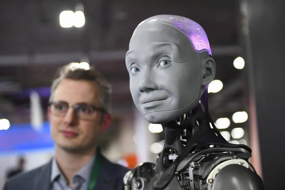 Robot humanoide de aspecto realista con luz púrpura en la cabeza, observado por un hombre con gafas en una feria tecnológica.