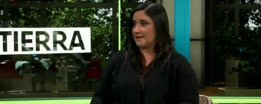 Mujer de cabello negro sentada en un estudio de televisión con el rótulo 'Tierra' visible al fondo.
