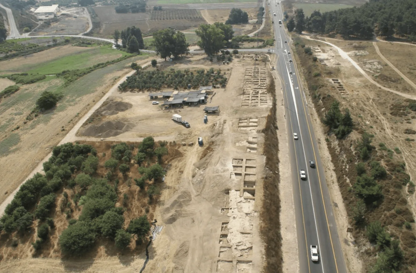 Vista aérea de un extenso sitio arqueológico con estructuras excavadas a lo largo de una carretera de doble sentido, rodeado de campos y árboles.