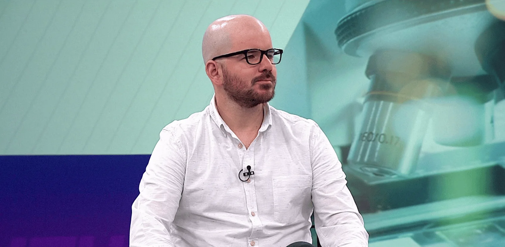 Hombre calvo con gafas y camisa blanca, con micrófono de solapa, sentado en un estudio de televisión con fondo verde y microscopio.