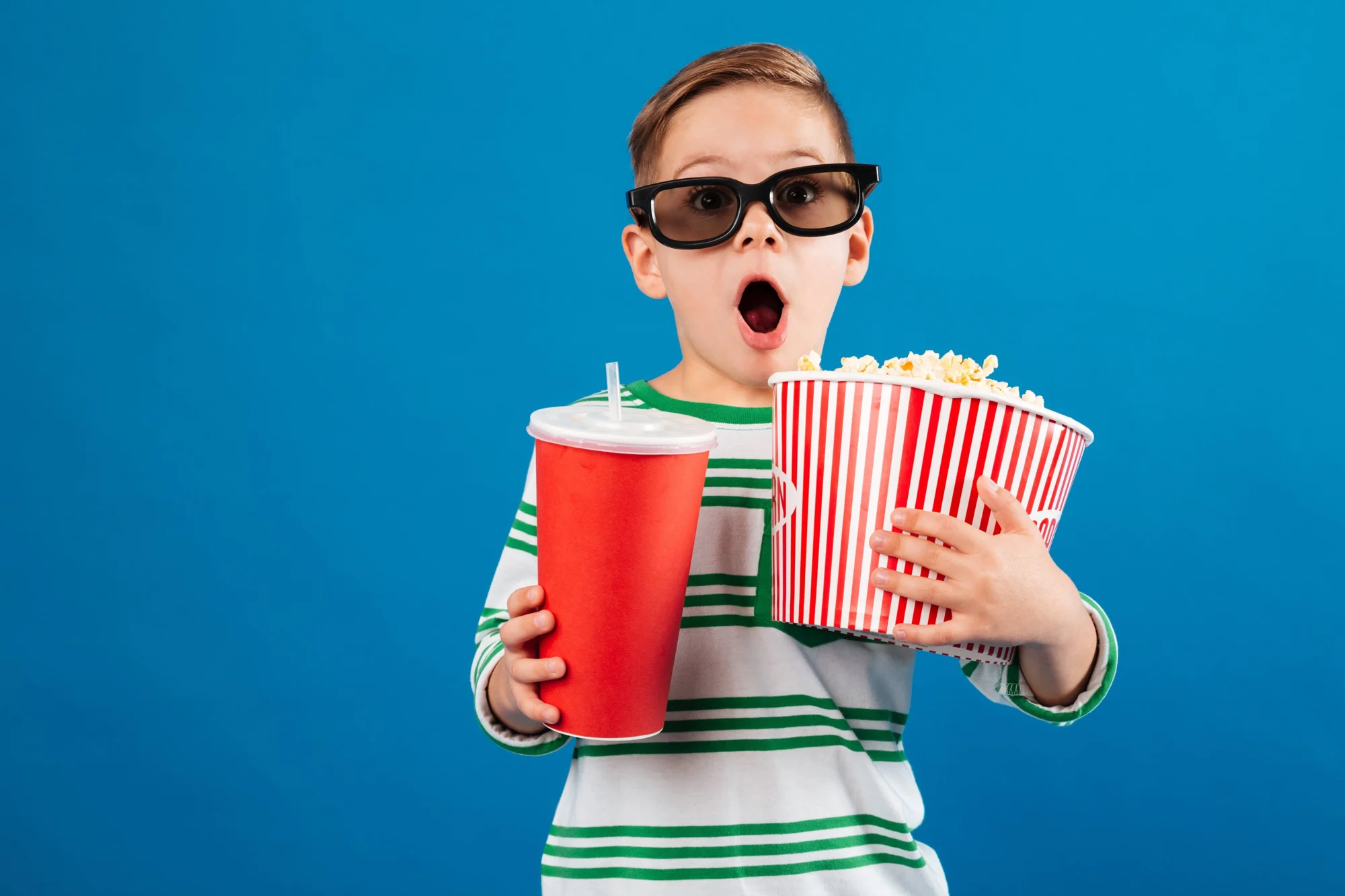 Niño con lentes 3D y boca abierta sostiene un cubo de palomitas y una bebida roja, expresando sorpresa.