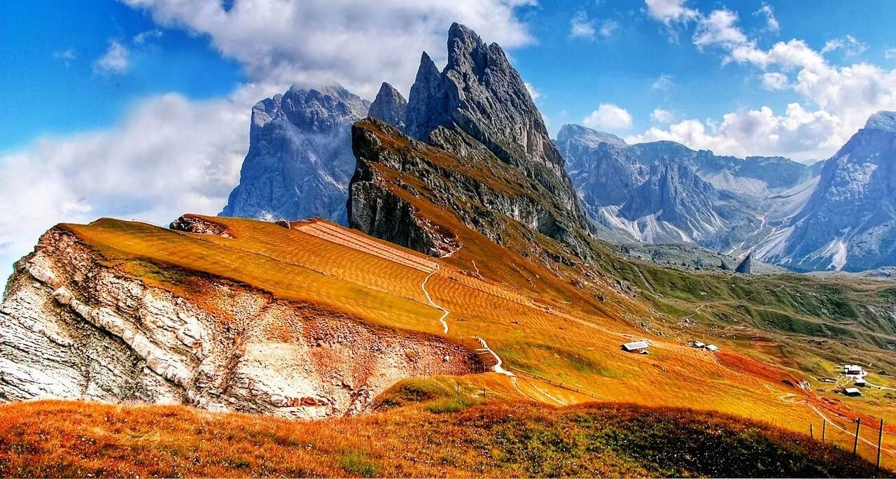 Paisaje alpino con praderas doradas otoñales, senderos de montaña, pequeñas cabañas y picos rocosos escarpados bajo cielo azul con nubes.