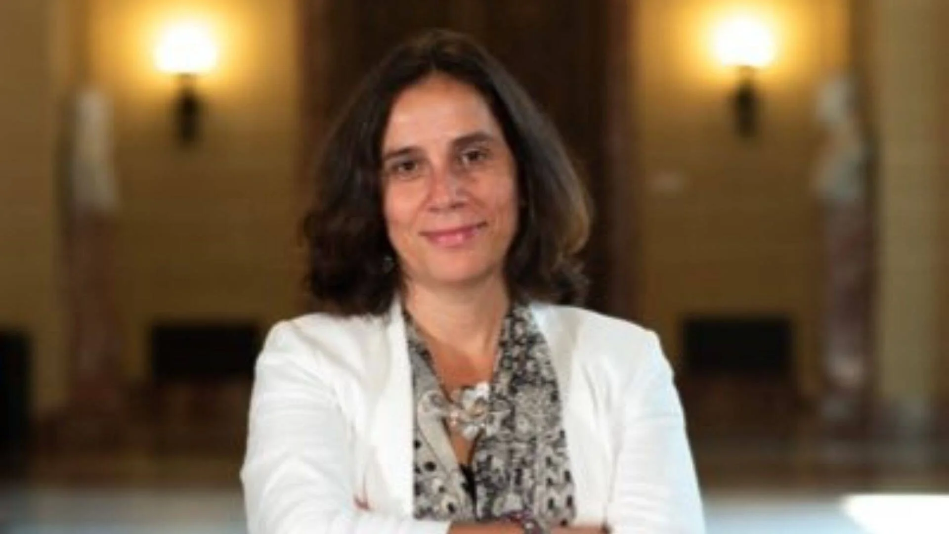 Mujer de cabello oscuro con blazer blanco y blusa estampada, posando con brazos cruzados en un salón de edificio institucional.