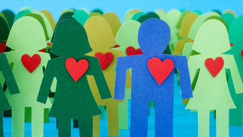 Grupo de figuras humanas recortadas en papel de colores verde y azul, cada una con un corazón rojo, tomadas de la mano.