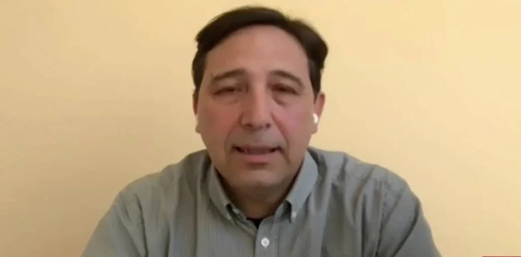 Hombre de mediana edad con camisa gris y auricular en el oído habla frente a la cámara en una videollamada.