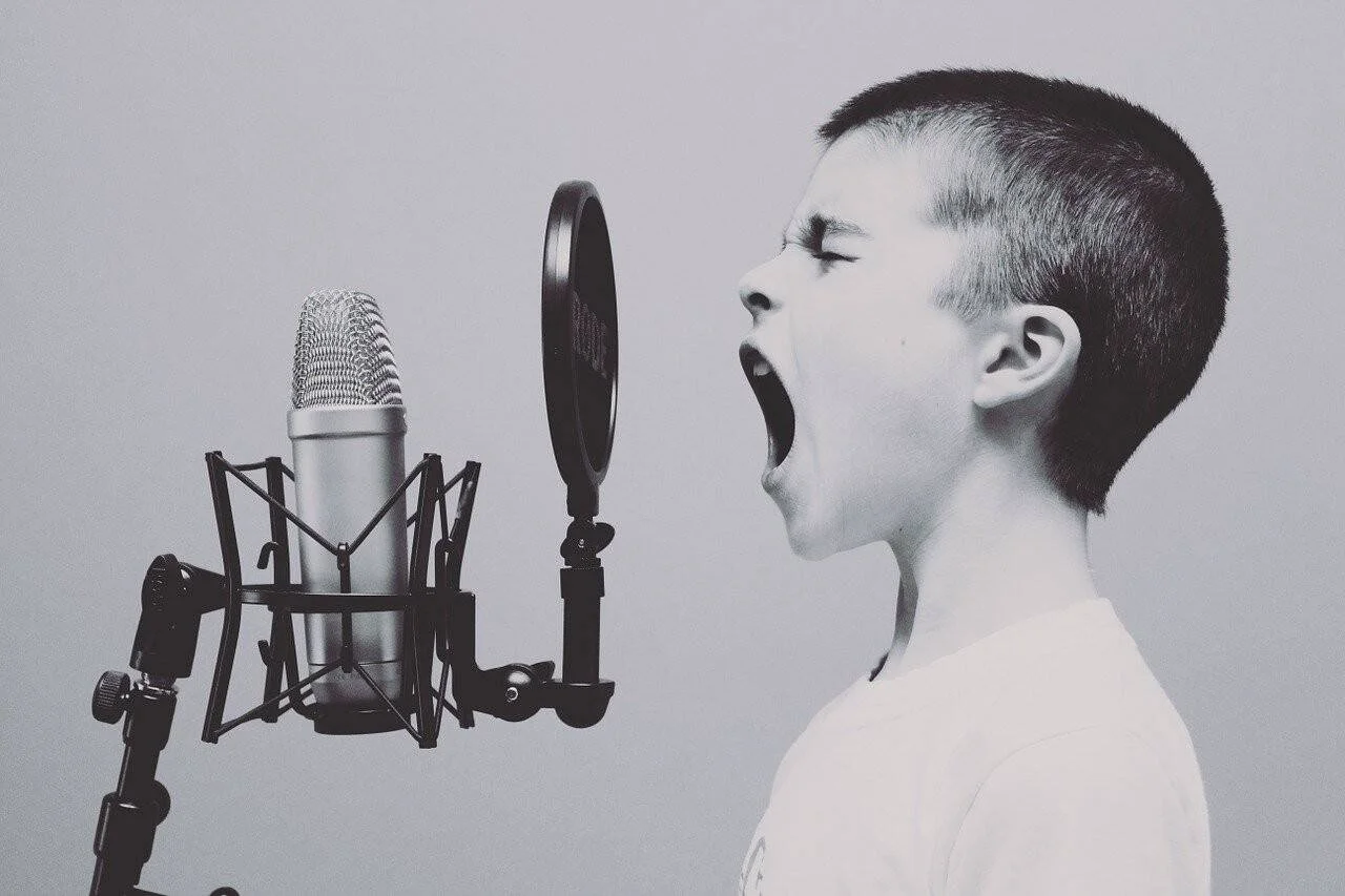 Niño con la boca abierta cantando ante un micrófono de condensador con filtro antipop, en blanco y negro.