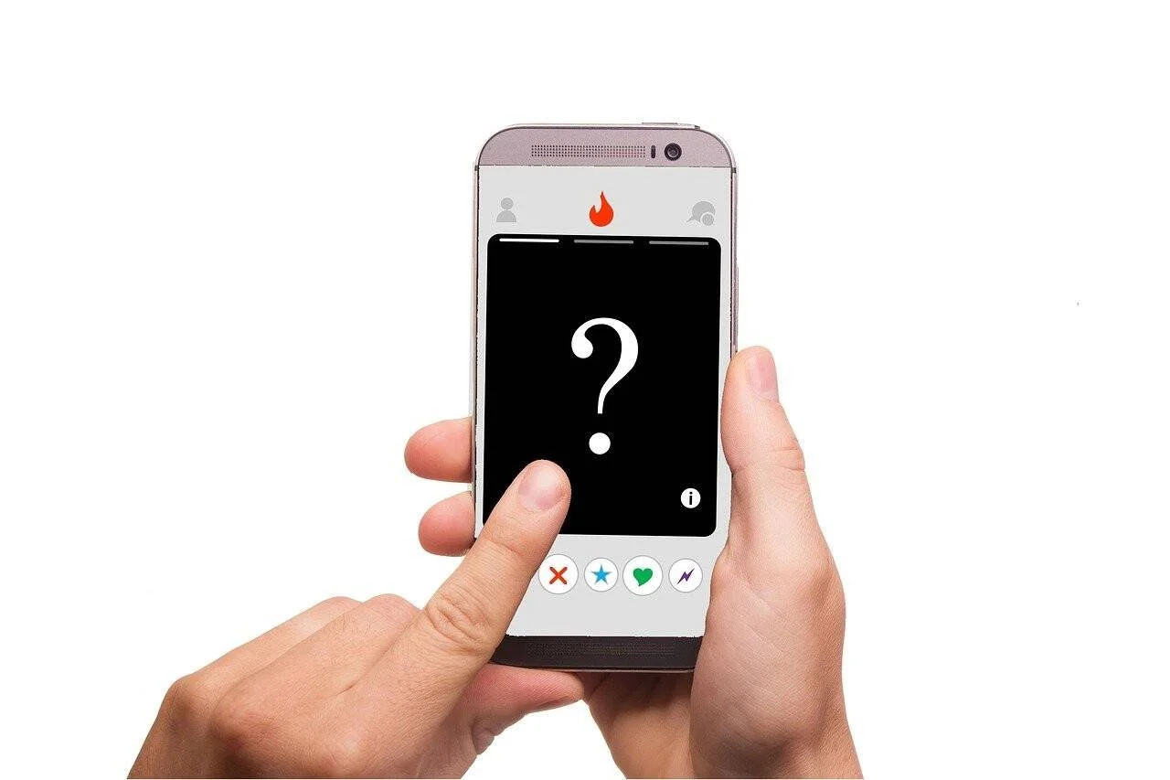 Manos sosteniendo un smartphone con la app Tinder abierta, mostrando un perfil anónimo con signo de interrogación en pantalla.