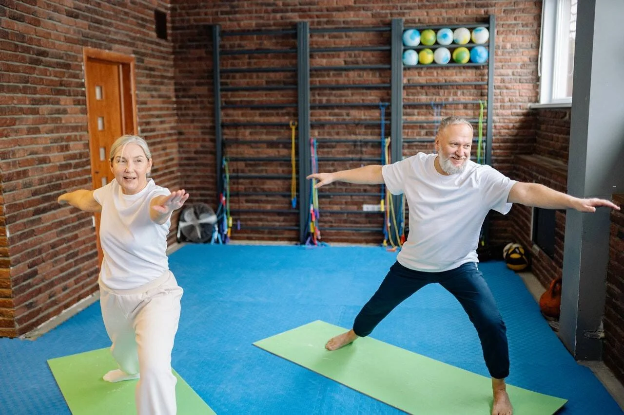 Dos personas mayores sonrientes realizan la postura del guerrero sobre colchonetas en un gimnasio con paredes de ladrillo.