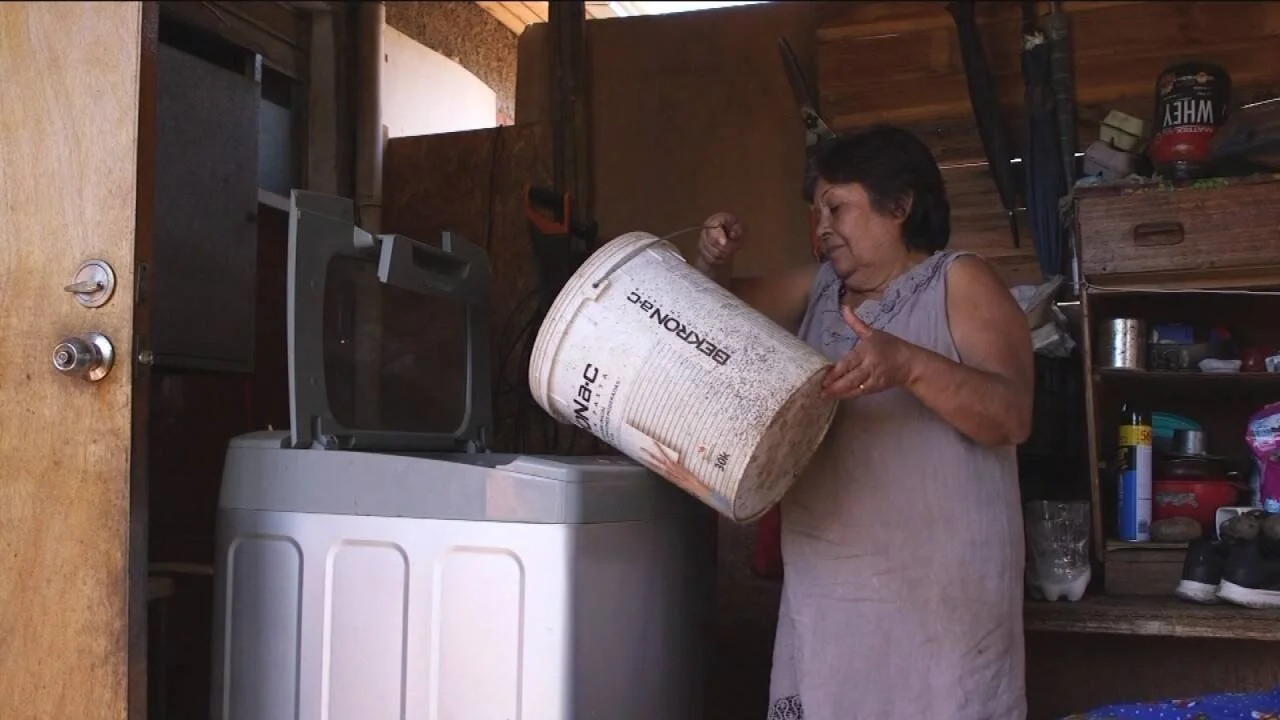 Una mujer vierte agua desde un balde industrial hacia un lavarropas, evidenciando falta de suministro de agua corriente.