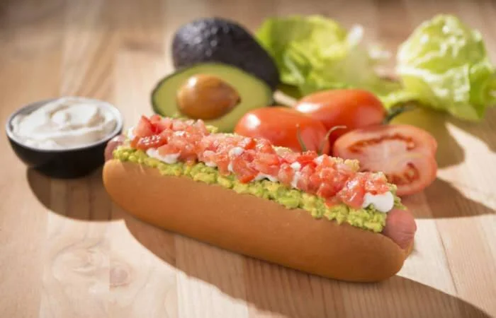 Hot dog en pan con guacamole y tomate picado, acompañado de crema agria, aguacate, tomate y lechuga al fondo.