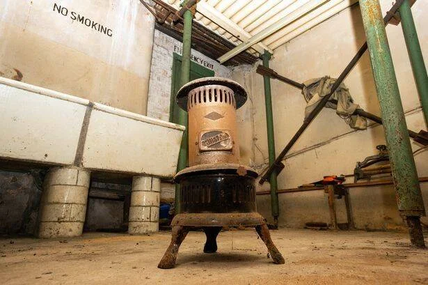 Vieja estufa de queroseno oxidada sobre patas metálicas en un espacio industrial deteriorado con cartel de prohibido fumar al fondo.