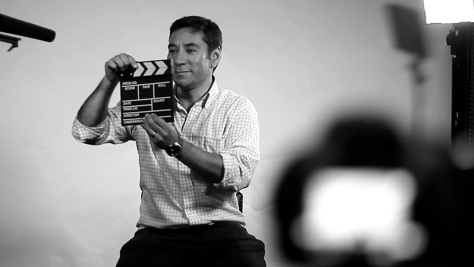 Hombre con camisa a cuadros sostiene una claqueta de cine frente a una cámara en un set de grabación, en blanco y negro.