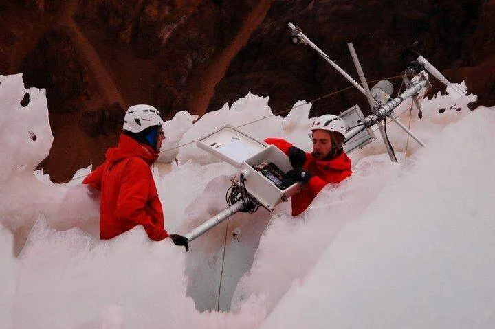 Dos técnicos con cascos y ropa roja instalan equipos de monitoreo entre formaciones de hielo en un glaciar.