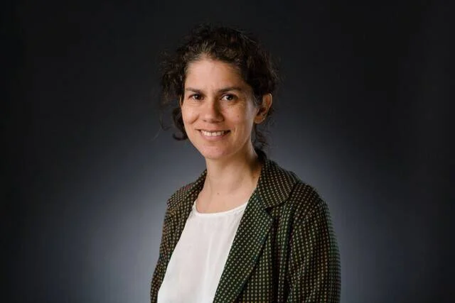 Mujer de cabello rizado recogido, con blazer verde oscuro y blusa blanca, posando ante fondo gris oscuro.