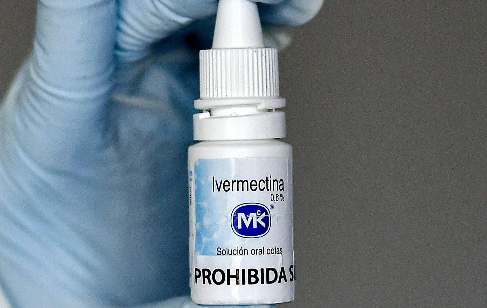 Mano con guante azul sostiene un frasco de ivermectina 0,6% en solución oral gotas, con etiqueta que indica 'Prohibida su venta'.