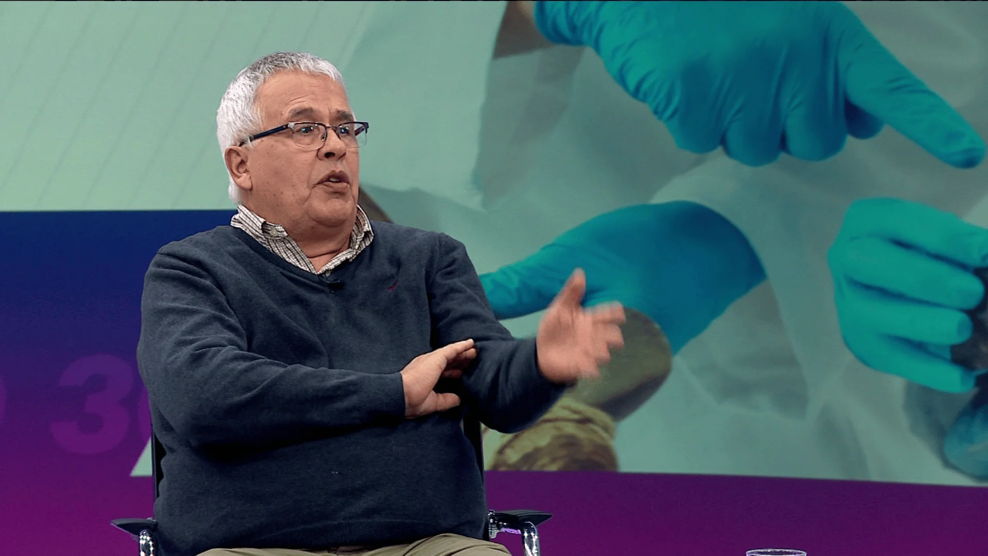 Hombre de cabello blanco y gafas gesticula mientras habla en un estudio de televisión con pantalla médica al fondo.