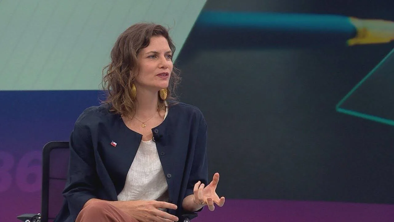 Mujer con chaqueta azul marino y pin de la bandera chilena habla gesticulando en un estudio de televisión.
