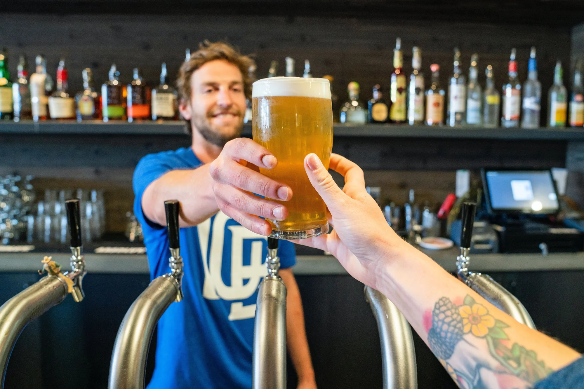 Un bartender sonriente entrega un vaso de cerveza rubia con espuma a un cliente con brazo tatuado, frente a grifos de barril.