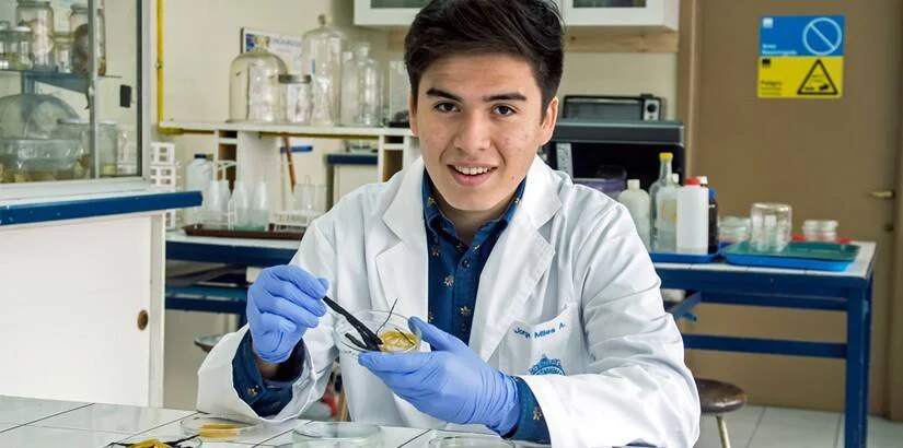 Joven con bata blanca y guantes azules manipula una muestra con pinzas en un laboratorio científico.