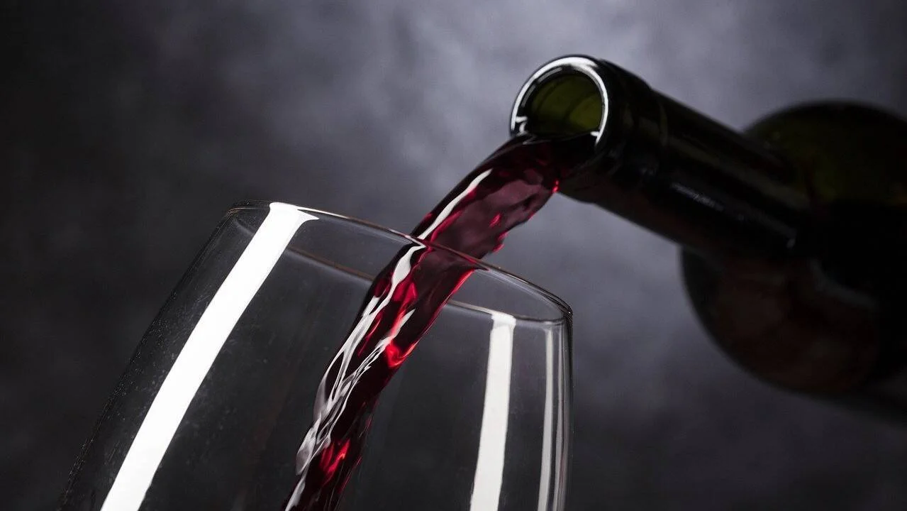 Vino tinto siendo vertido desde una botella oscura hacia una copa de cristal transparente sobre fondo gris.