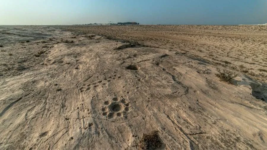 Superficie rocosa en desierto árido con grabados circulares antiguos y edificaciones al fondo en el horizonte.