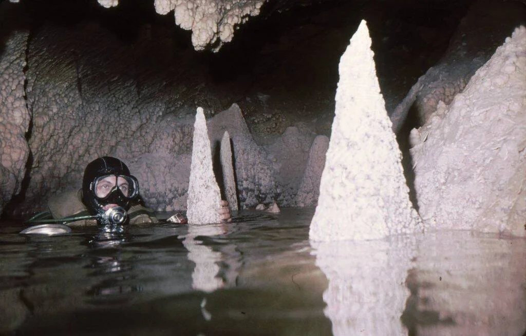 Un buzo equipado emerge entre estalagmitas blancas en un lago dentro de una cueva subterránea con estalactitas en el techo.